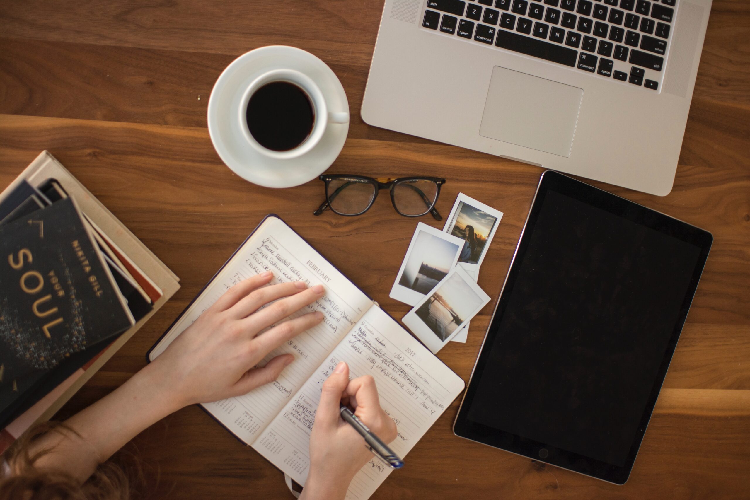 Photo of hands writing in a journal near a computer and coffee and photos