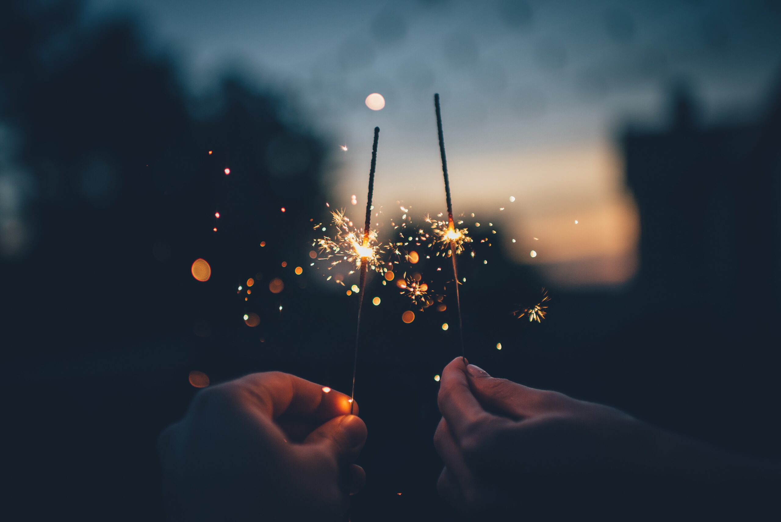 Dark photo of two hands holding sparklers with sunset in the background.